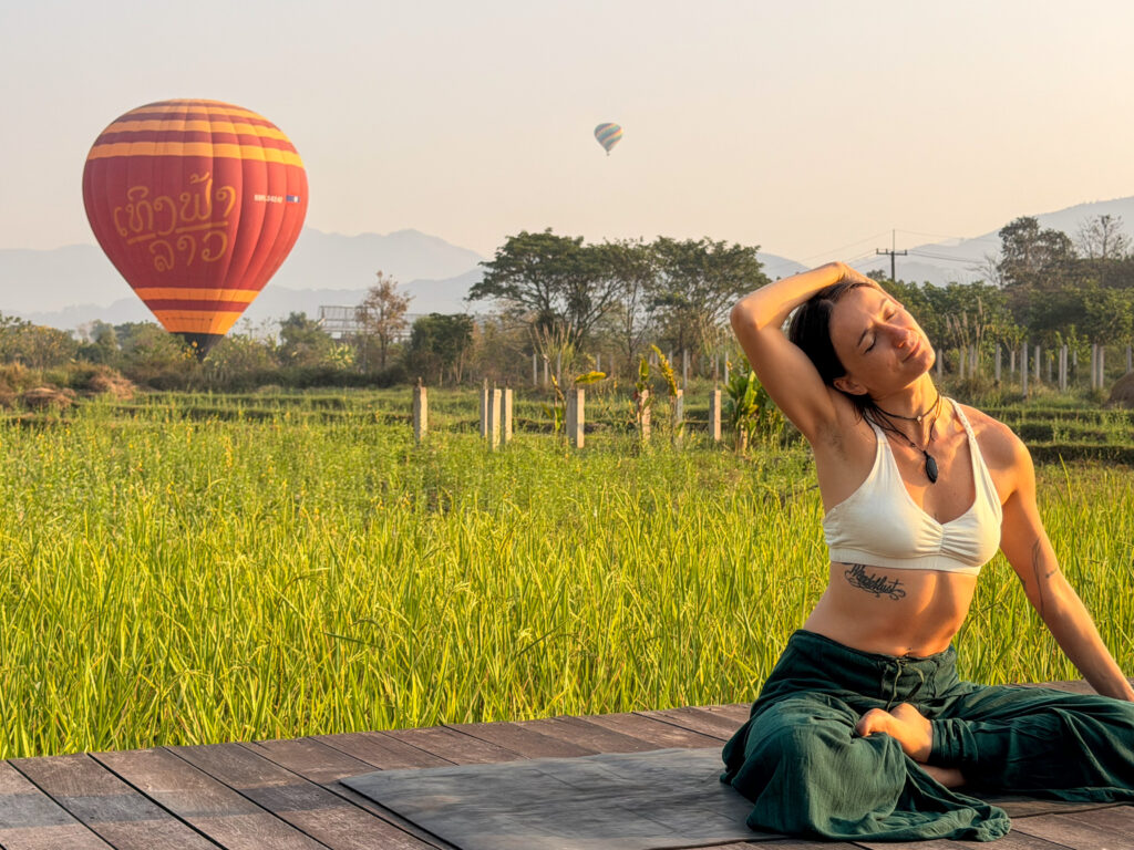 A yoga teacher holds her hand over her head, tilting it to the side for a neck stretch, sitting on a wooden platform with a green field and hot air balloons behind her